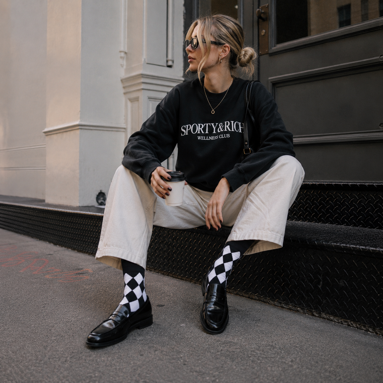 Person wearing a black 'Sporty & Rich' sweatshirt sitting on a step.