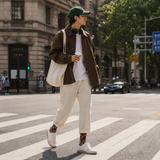Person crossing a street wearing a brown jacket, white pants, and a green cap.