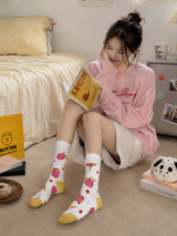 Woman in pink hoodie and patterned socks sitting on the floor with a book in a bedroom.