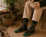 Person wearing patterned socks sitting on a wooden chair with a cup, surrounded by plants and a basket.